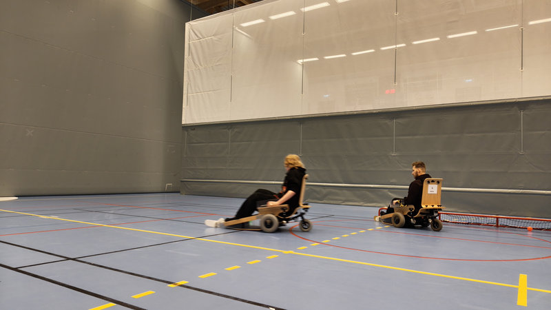 To el-innebandyspillere i rullestolkjelker under trening i Trønderhallen. En spiller i bevegelse med ball, og en annen spiller med utstyr ved sidelinjen.